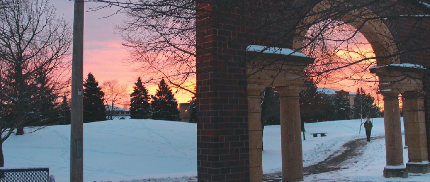 A student walking outside on the campus premises towards the arch on a winter evening with the sunset in the background