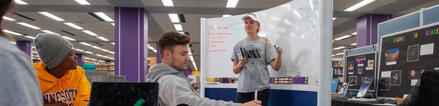 One student, standing at a whiteboard, working with three other students, sitting at a table taking notes