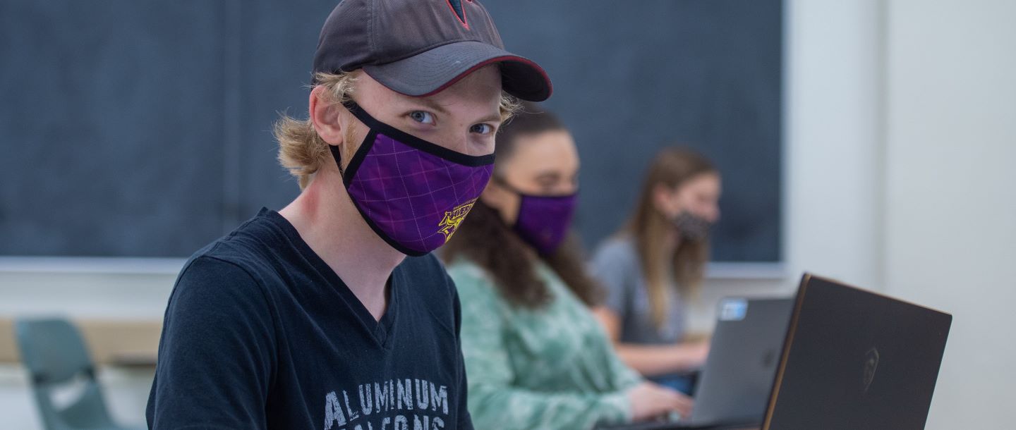 A student using his laptop in a classroom with his maverick cloth mask on and other students in the background